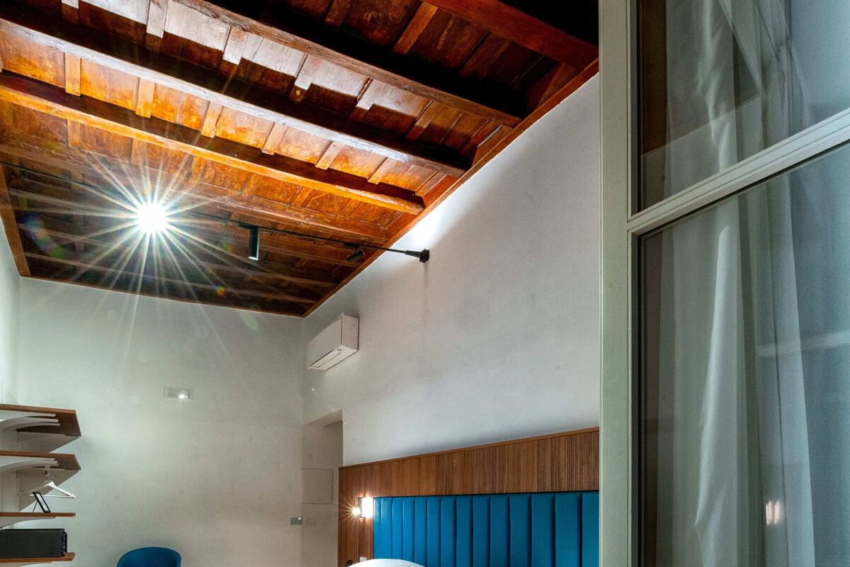 Un dormitorio moderno con techo de madera, una cama con cabecera azul, un ventanal con cortinas y un escritorio con silla.