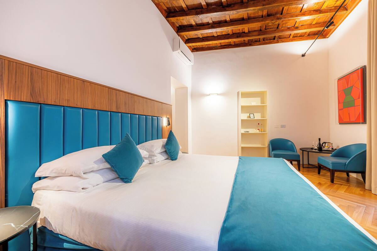 Habitación de hotel con techo de madera, cama con ropa de cama azul, una mesita, dos sillas y una estantería.