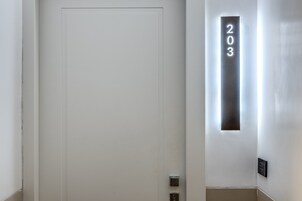 Egy fehér ajtó, rajta egy „203”-as számú táblával, és a falra szerelt lámpatesttel.