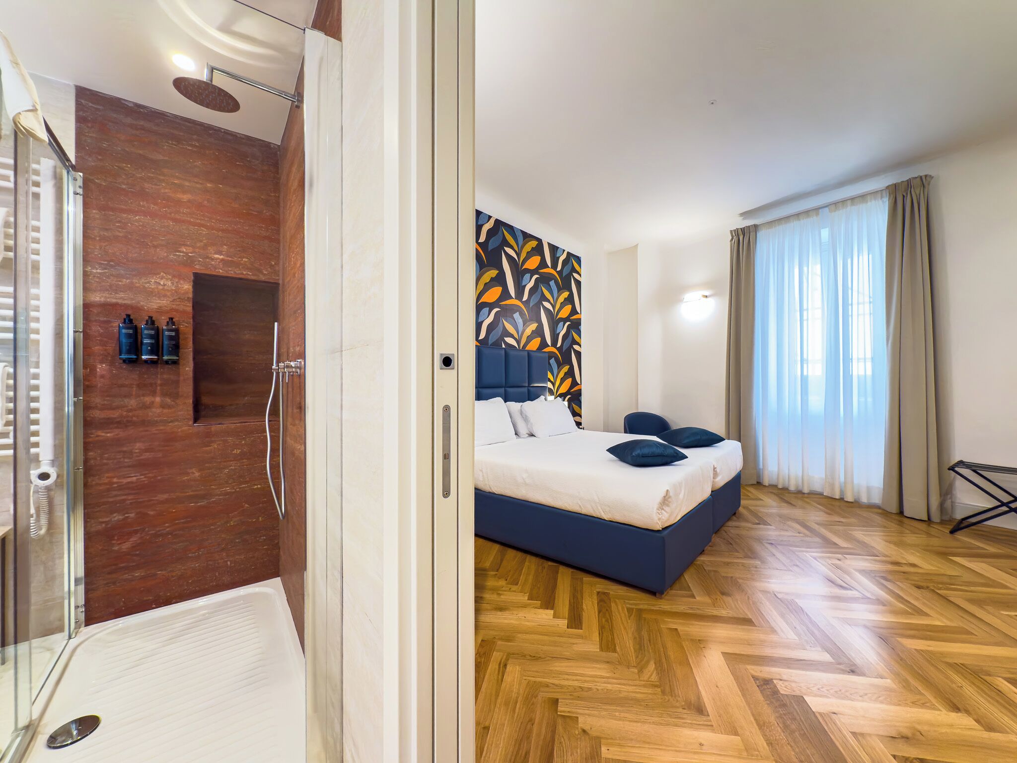 A modern hotel room with a wooden floor, a large bed with blue bedding, a shower area, and a wall with a colorful abstract design.
