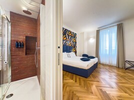 A modern hotel room with a wooden floor, a large bed with blue bedding, a shower area, and a wall with a colorful abstract design.