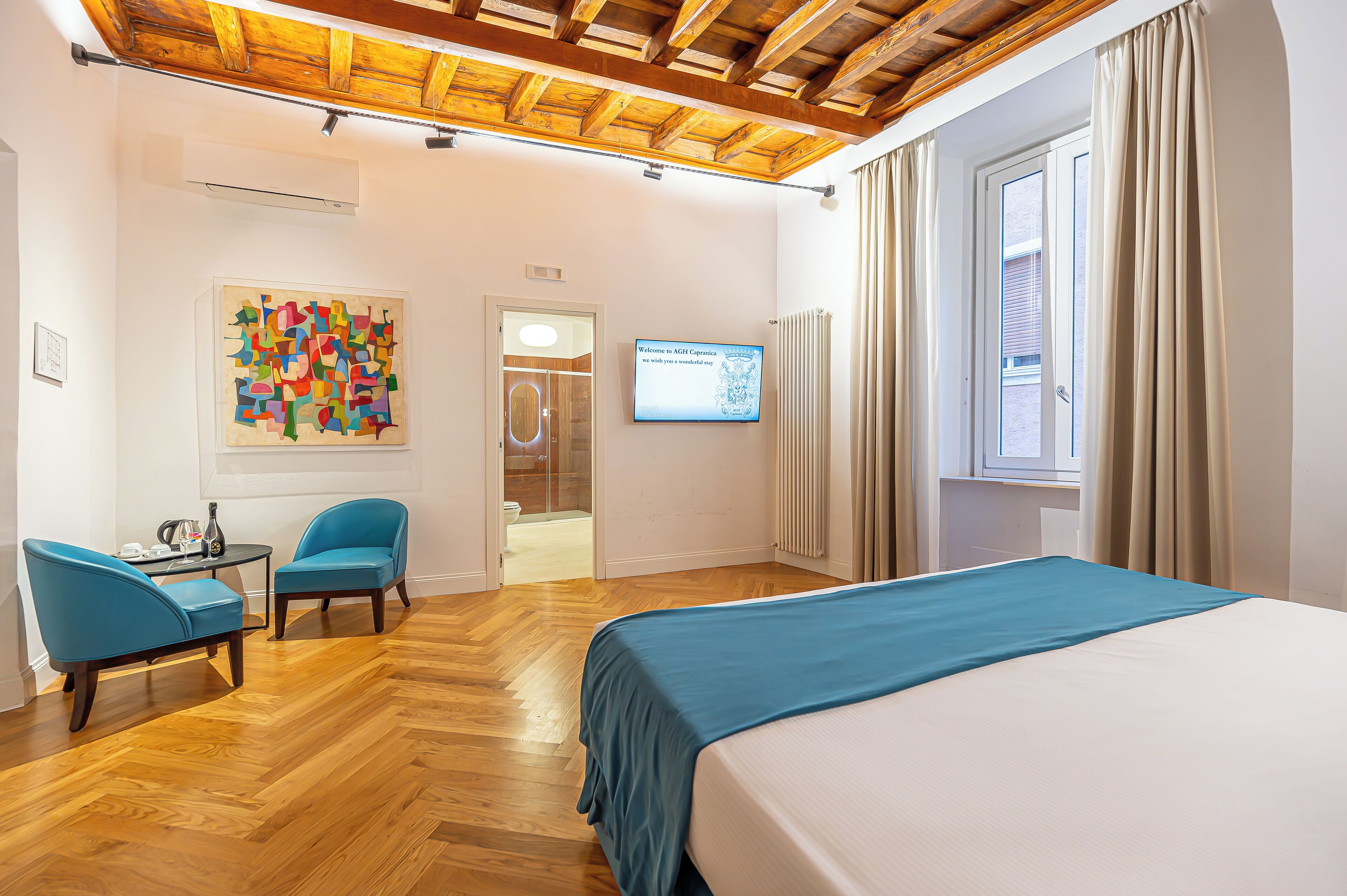 A hotel room with a wooden ceiling, a bed with a blue bedspread, two blue chairs, a small table with a vase, a colorful abstract painting, and a television screen.