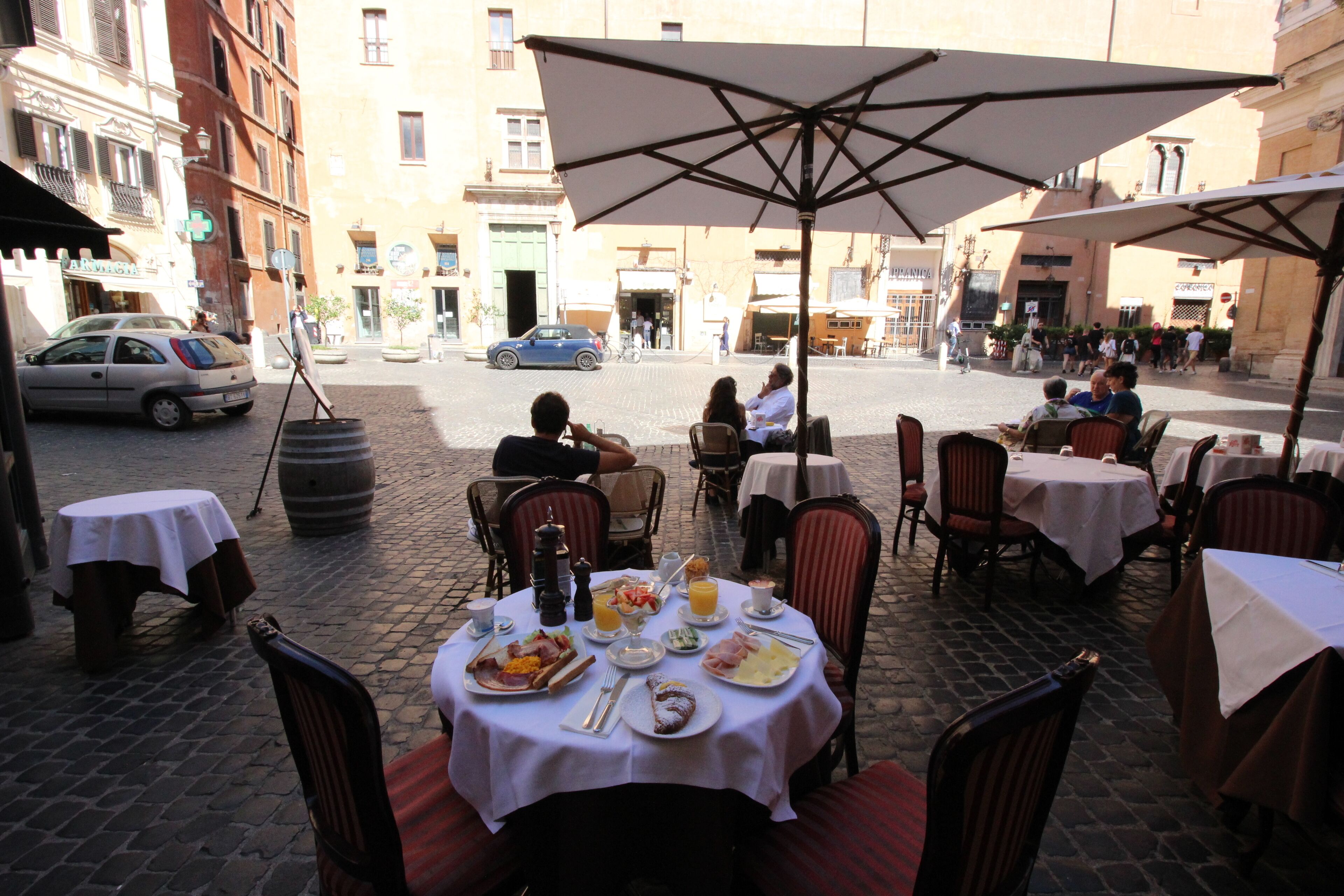Un café al aire libre con mesas y sillas, un paraguas grande y una calle adoquinada al fondo.