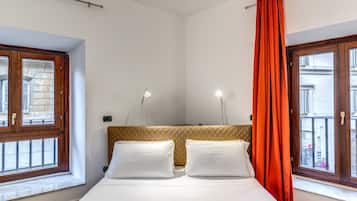 A hotel room with a bed, two bedside tables with lamps, a window with curtains, and a view of the street outside.
