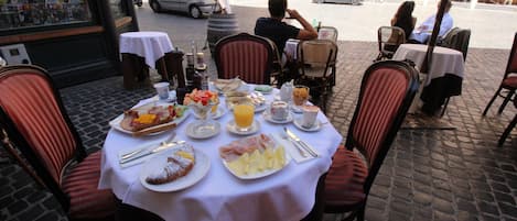 An outdoor cafe with tables set for breakfast, featuring plates of food, drinks, and a street view in the background.