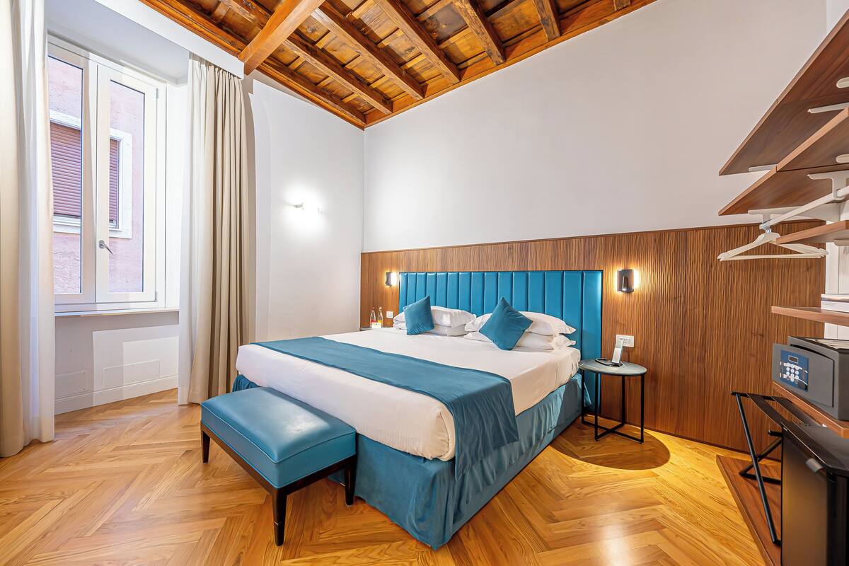Un dormitorio con techo de madera, una cama con ropa de cama azul, una mesita de noche y un escritorio pequeño con un ordenador.