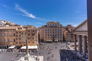 A historic square with classical architecture, including a fountain and columns, surrounded by buildings with balconies and outdoor seating.