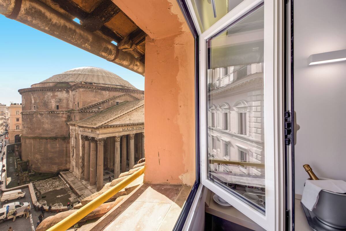 Una habitación con vista a un edificio antiguo, una ventana con una botella y una barandilla amarilla.