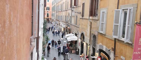 Un carrer estret flanquejat per edificis, alguns amb persianes. Hi ha gent caminant i d'altres de peu, i algunes taules i caires davant d'un edifici.