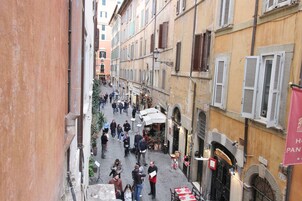 A narrow street with buildings on either side, some with shutters. There are people walking and some standing, and a few tables and chairs outside a building.
