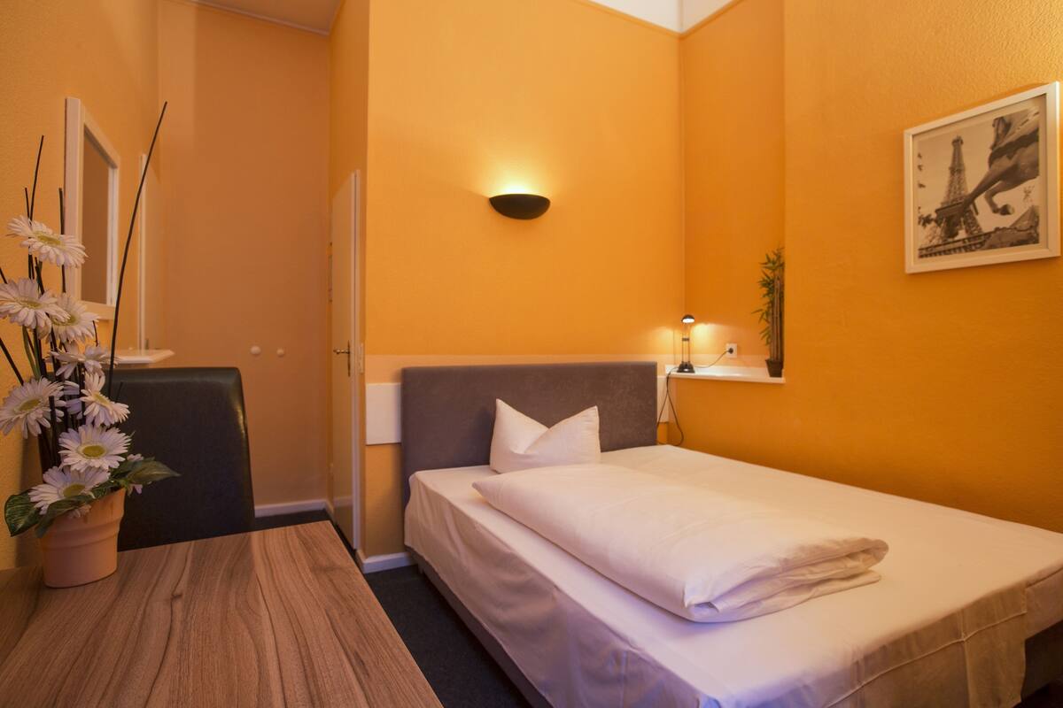 A hotel room with a bed, a desk, a chair, a vase with flowers, and a wall-mounted light fixture.
