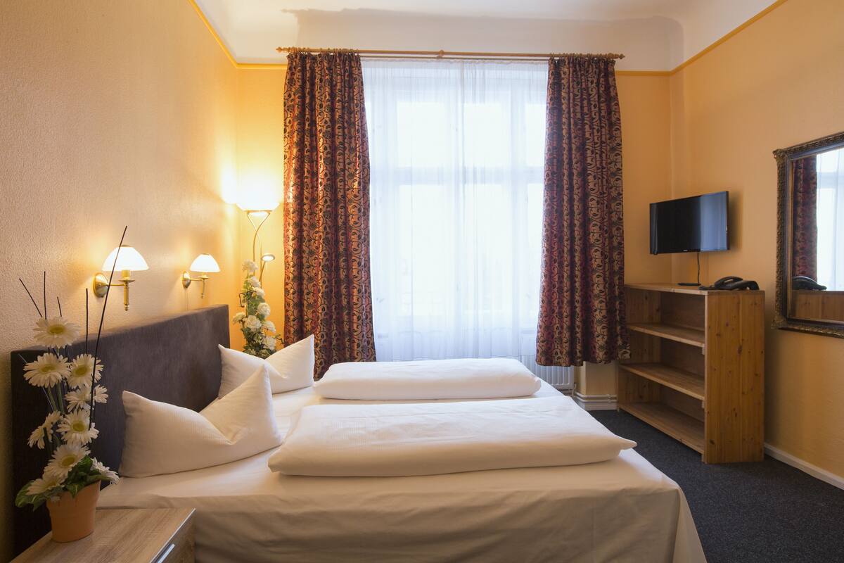 A hotel room with a bed, a TV, a wooden shelf, and a window with curtains.