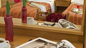 Une chambre d’hôtel avec un lit, une table de chevet, une lampe et un miroir qui reflète la pièce. Sur la table, on trouve un vase avec des fleurs, une tasse et un magazine.