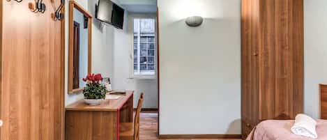 Una camera da letto con un'armadio in legno, una scrivania con una pianta, un letto con una coperta rosa e una televisione a parete.
