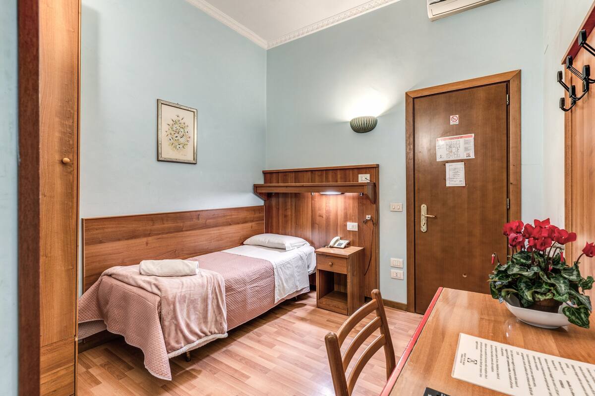 A small, neatly arranged room with two single beds, a wooden nightstand, a small table with a plant, and a door with a sign.