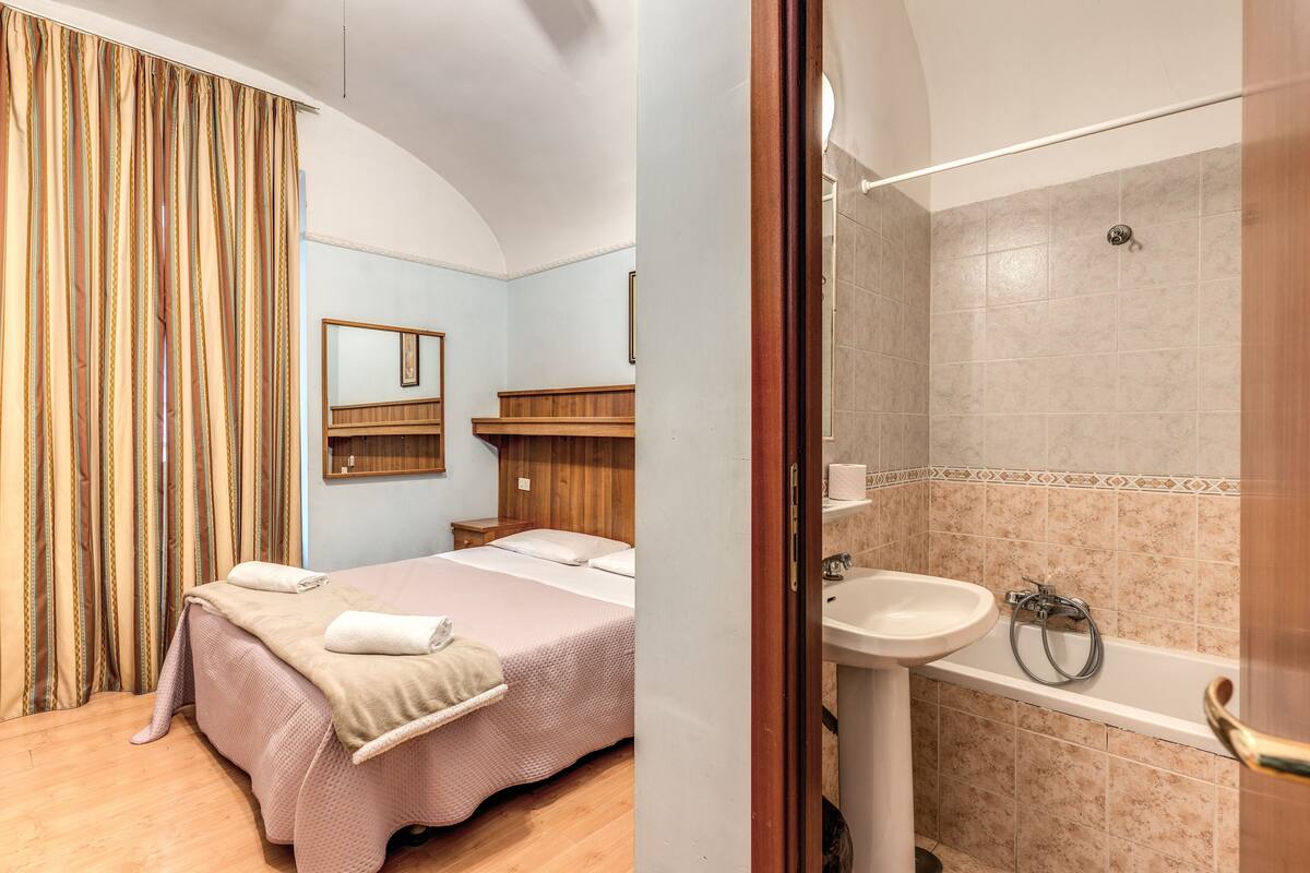 A bedroom with a bed, a wooden dresser, and a mirror. The bathroom has a sink and a shower.