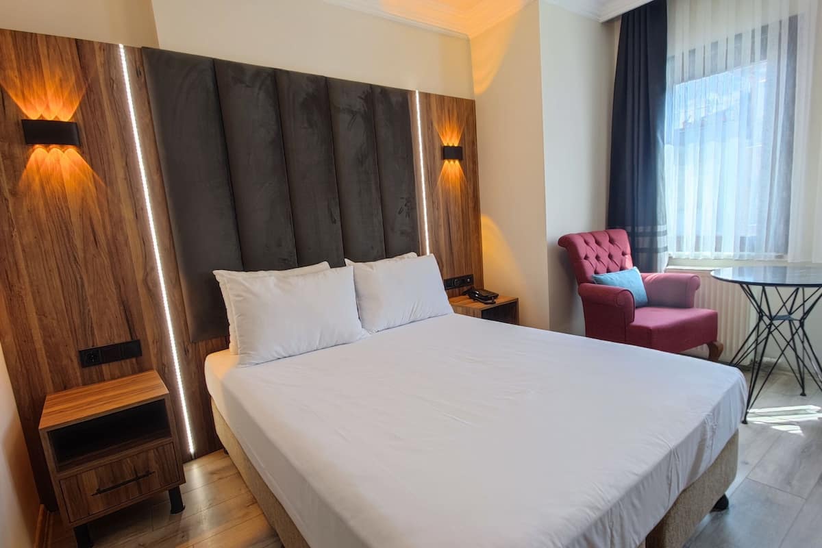 A modern hotel room with a large bed, wooden headboard, bedside tables, a red armchair, and a small table with a chair.