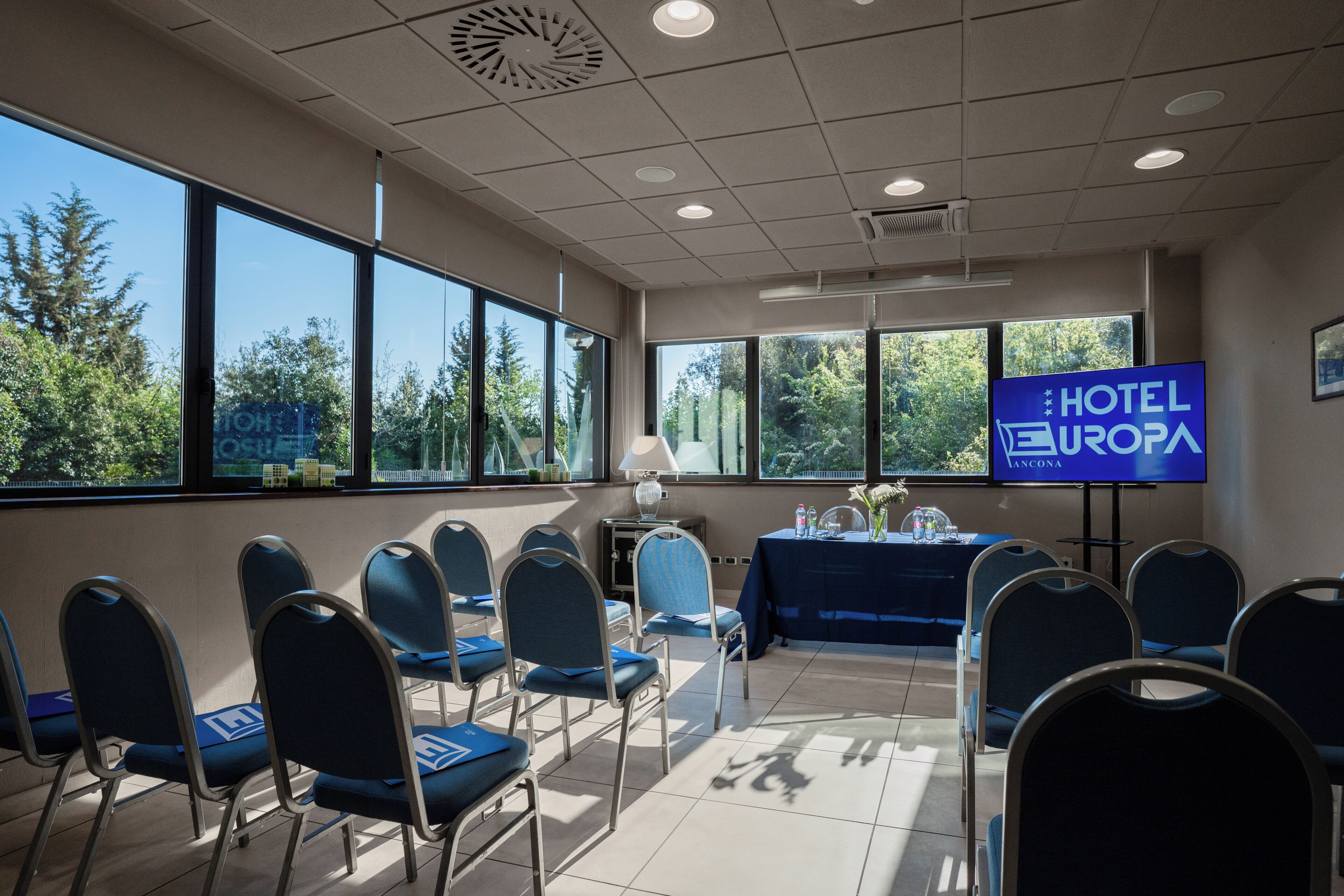 Un salón de conferencias con una pantalla grande mostrando 'HOTEL EUROPA LANCONA', sillas dispuestas en filas y ventanas amplias con vistas a árboles.