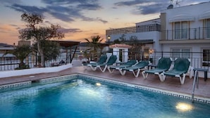 A swimming pool with lounge chairs, a pergola, and a view of a cityscape at sunset. - Hotel Ninays (Lloret de Mar)