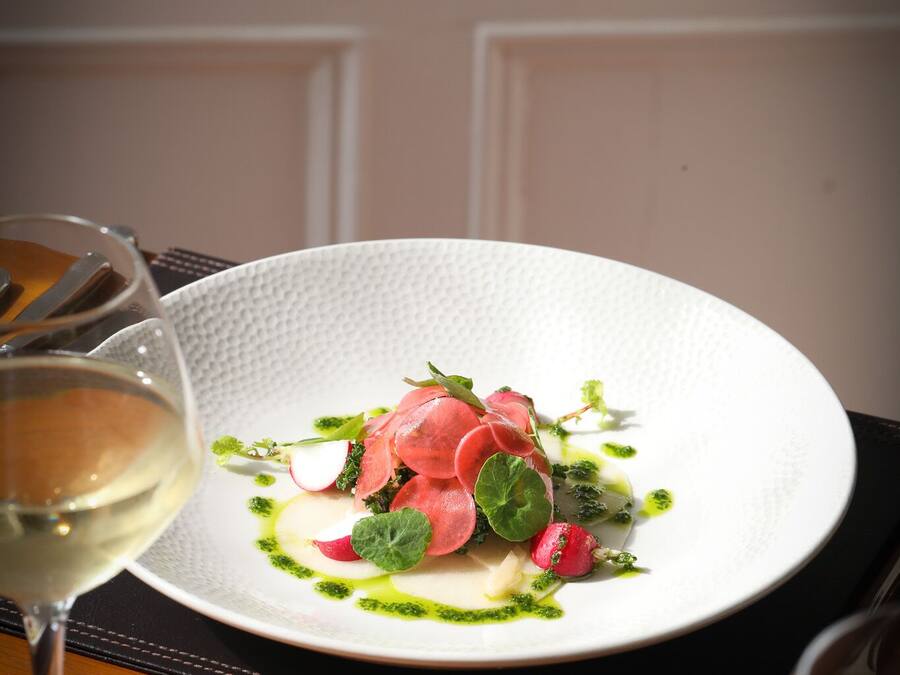A white plate with a dish of food, two glasses of white wine, and a black placemat on a wooden table.