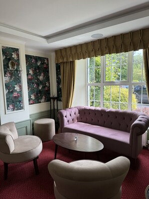 Una habitación con sofá capitonado, dos sillones, una mesita redonda, papel tapiz floral y una ventana con cortinas.