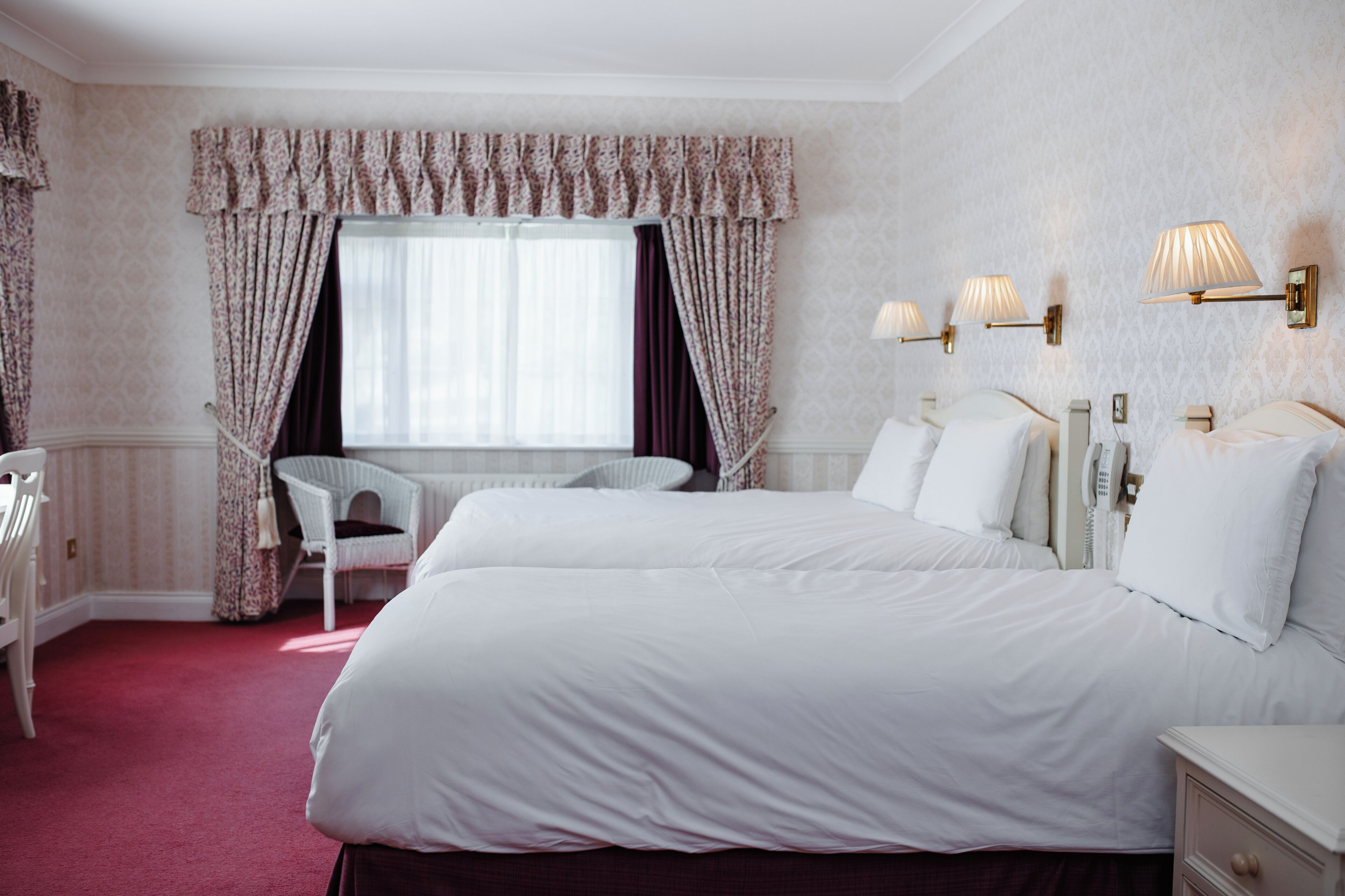 Ett hotellrum med två sängar, ett fönster med gardiner, en stol och väggmonterade lampor.