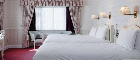 A hotel room with two beds, a window with curtains, a chair, and wall-mounted lamps.