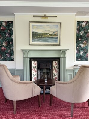 Una habitación con chimenea, dos sillones, una mesa con un vaso y papel tapiz floral.