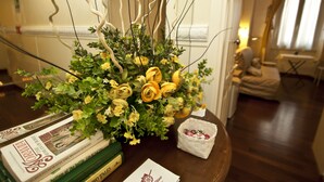 A room with a wooden table holding a bouquet of yellow roses, a basket of small items, and a stack of books. - Le Suite di Via Ottaviano (Rome)