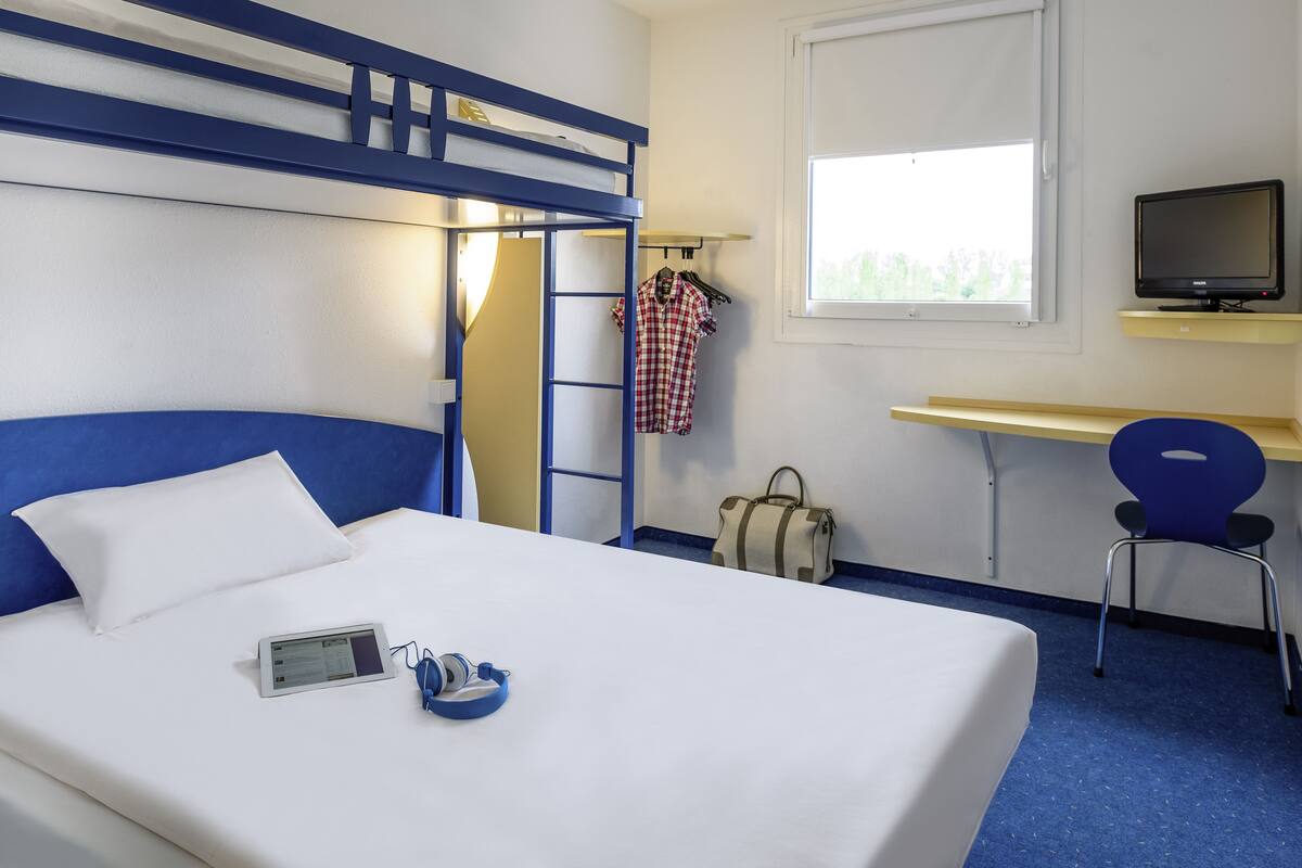 Habitación de hotel con una cama litera, un escritorio con una computadora, una silla, un televisor y una ventana.