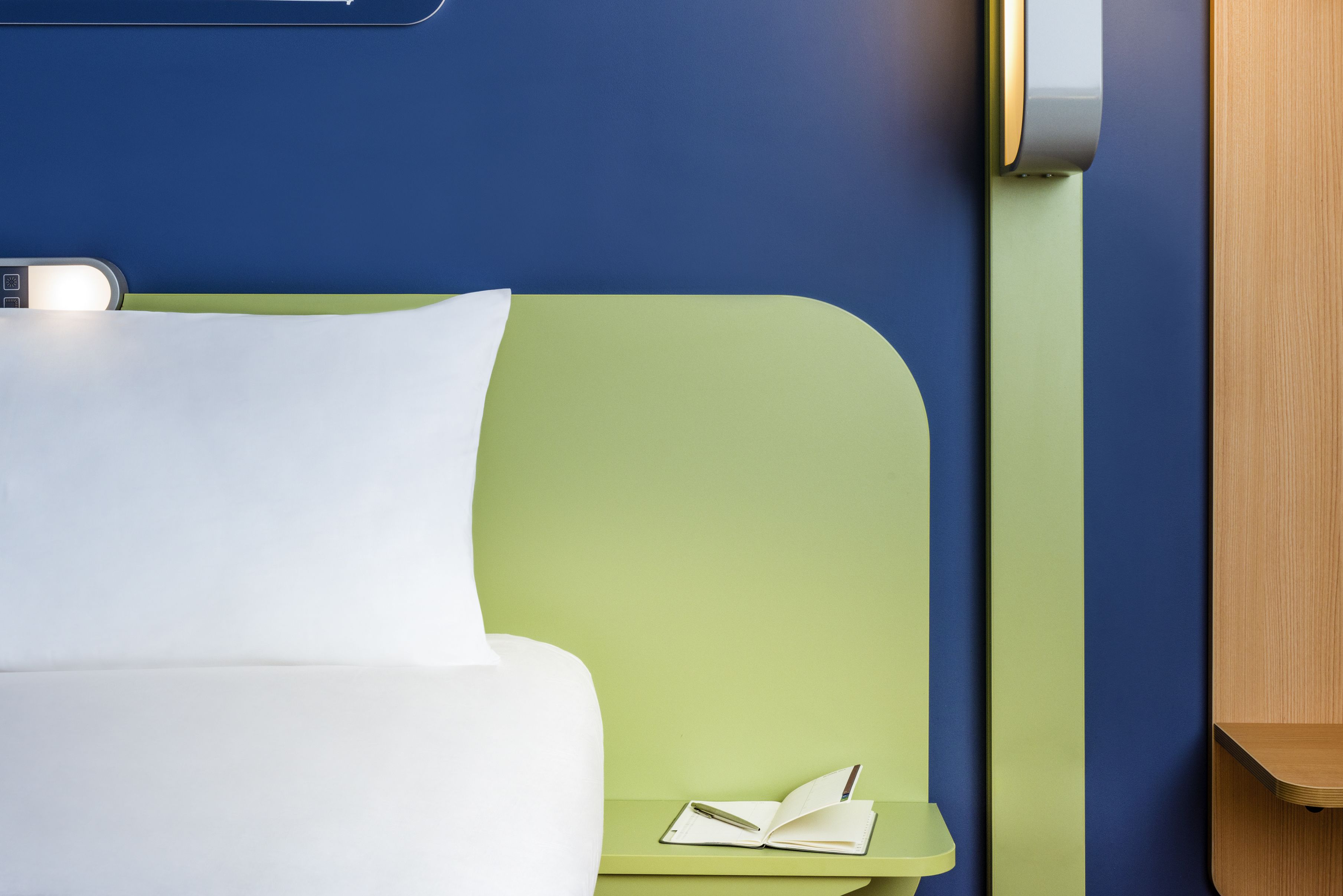 Una cama bien hecha con sábanas blancas, una mesita de noche verde con un libro abierto y un bolígrafo, y una pared azul con un panel de madera.
