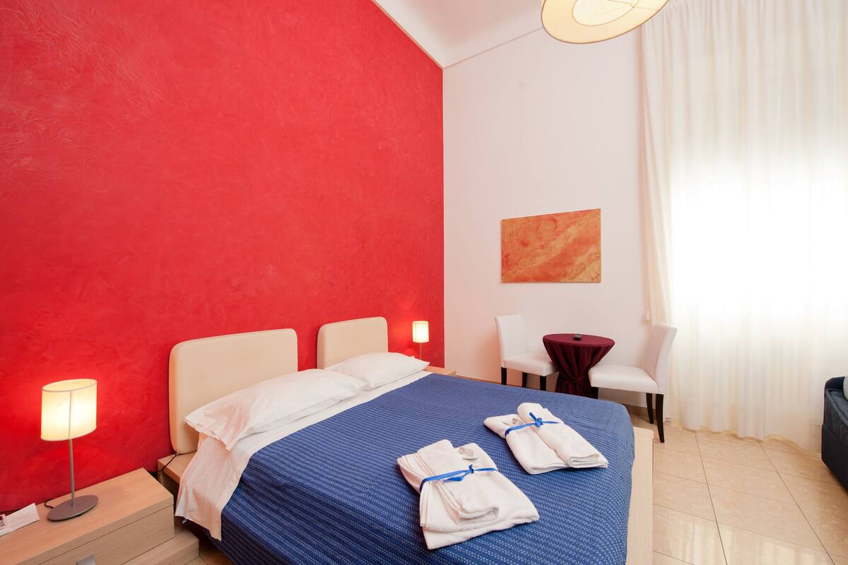 A hotel room with a red accent wall, a neatly made bed with blue bedding, a bedside lamp, a small table with a red tablecloth, and a chair.