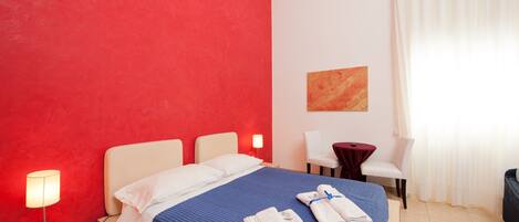 Una habitación de hotel con una pared de acento roja, una cama bien hecha con ropa de cama azul, una lámpara de noche, una mesita con mantel rojo y una silla.