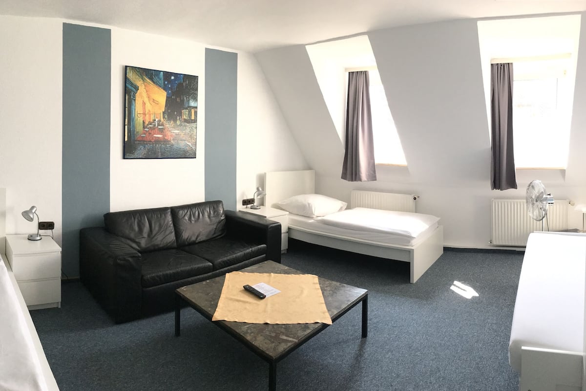 Une chambre d’hôtel avec un lit, un canapé, une petite table, un tableau au mur et une fenêtre avec des rideaux.