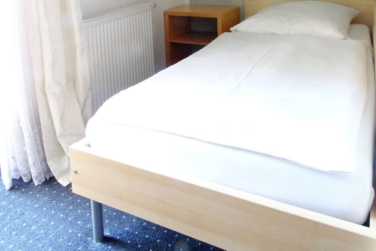 Un lit simple avec un cadre en bois et du linge de lit blanc, une petite table de chevet en bois, un radiateur et un tableau accroché au mur.