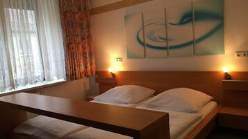 Ein Hotelzimmer mit Bett, Nachttischen, einer Holzbänklchen, einem Fenster mit durchsichtigen Vorhängen und einem blumigen Vorhang.
