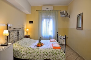 A hotel room with a bed, a bedside table with a lamp, a fruit basket on the bed, a TV mounted on the wall, and a window with curtains. - Le 4 Meraviglie (Rome)