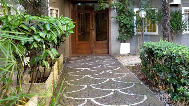 A pathway leading to a wooden door with a traditional design, flanked by lush greenery and a patterned brick floor.
