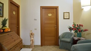 A room with two wooden doors, a statue, a chair, a small table with a vase of flowers, and a dresser with fruit on top.