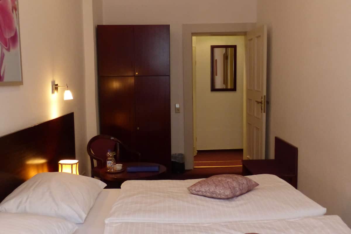 A hotel room with two beds, a nightstand, a lamp, a chair, and a door leading to another room.