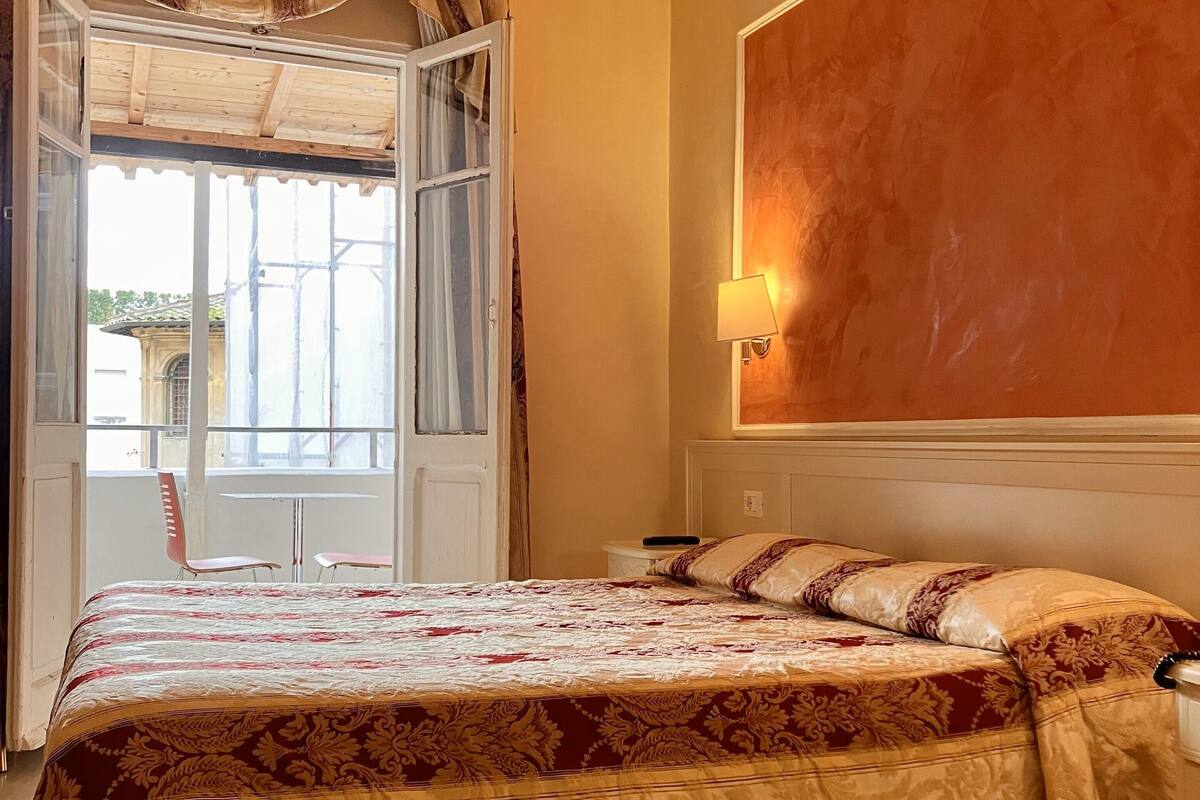 Une chambre à coucher avec un grand lit, une table de chevet, une lampe fixée au mur et une fenêtre avec des rideaux.
