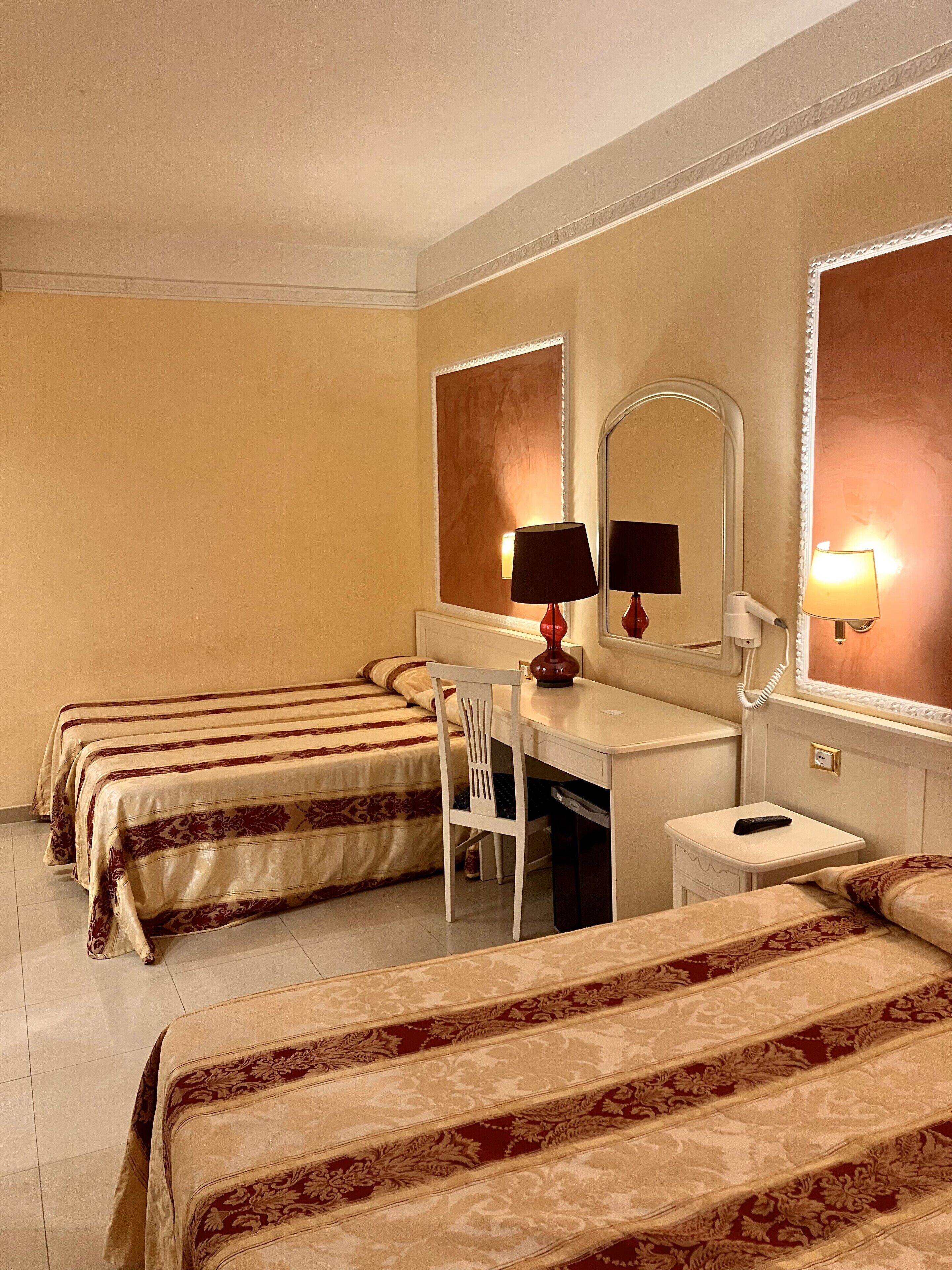 A hotel room with three beds, a desk, a chair, a mirror, and a nightstand with a lamp.