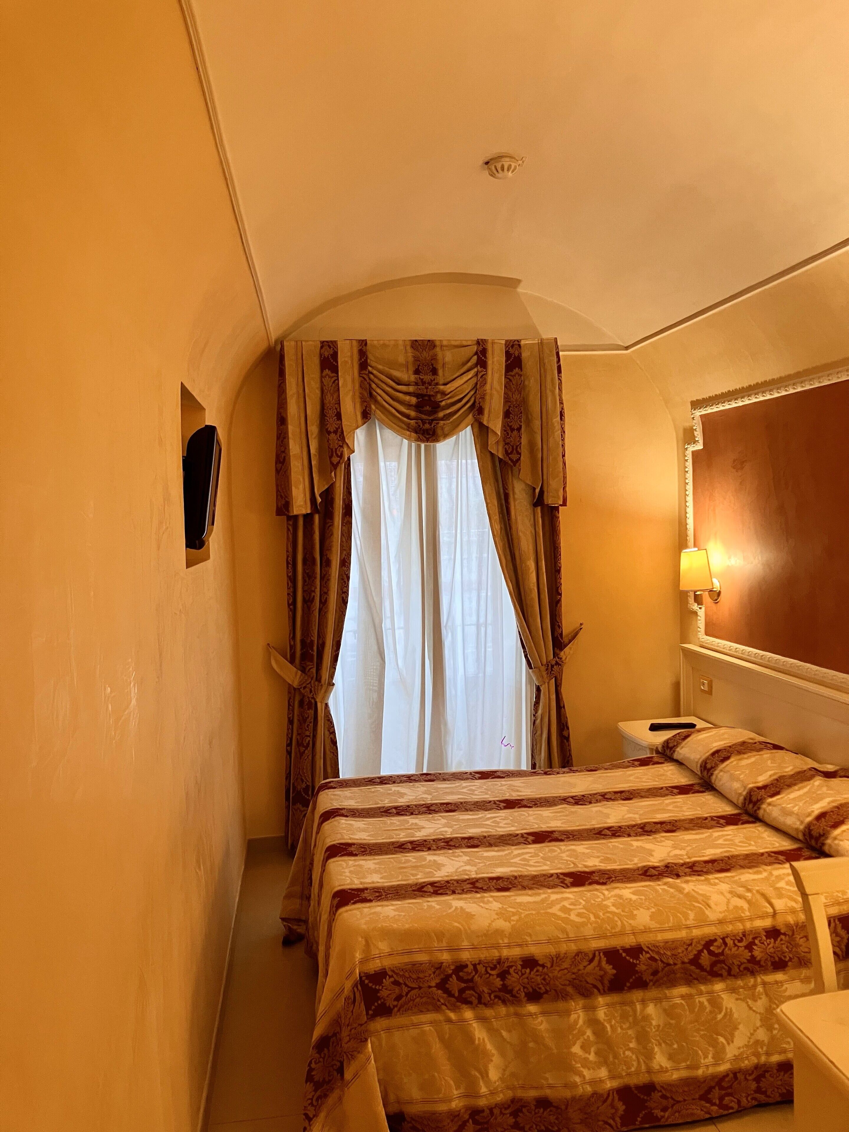Ein Hotelzimmer mit einem großen Bett, einem Nachttisch, einer Lampe und einem Fenster mit Vorhängen.