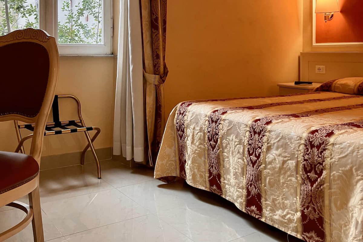 Une chambre d’hôtel avec un lit, une chaise, une table de chevet, une lampe et une fenêtre avec des rideaux.