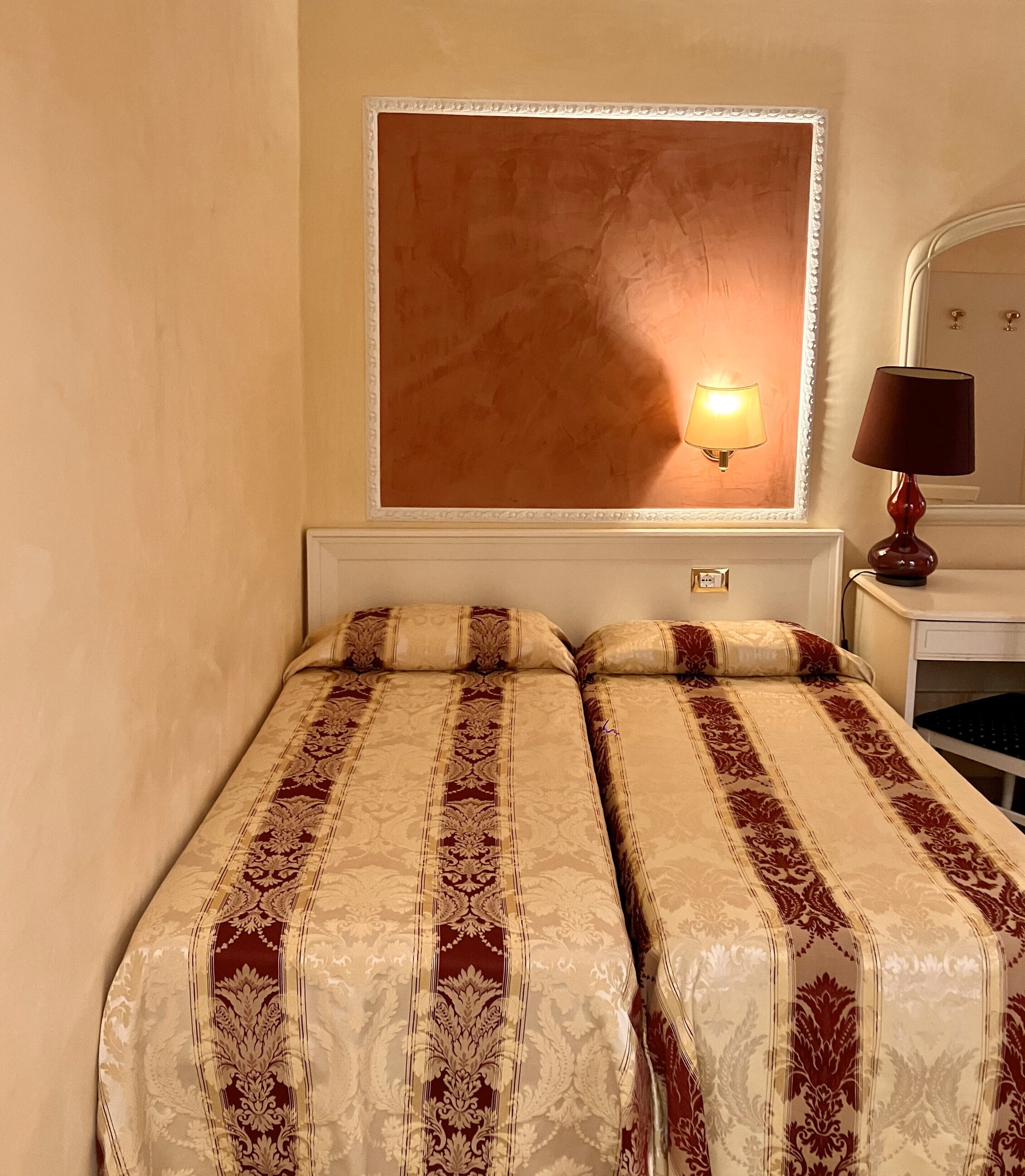 A hotel room with two beds, a nightstand, a lamp, and a wall-mounted light fixture.
