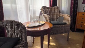 A room with wicker furniture, a wooden table with glasses and a bottle, and a view through sheer curtains. - Hotel Maison Am Olivaer Platz (Berlin)