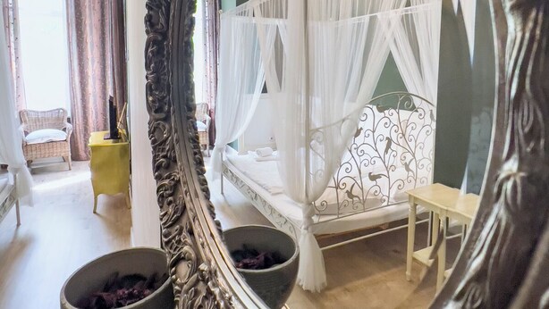 A bedroom with a four-poster bed, a large ornate mirror, and a view of a cityscape through the window. - Hotel Maison Am Olivaer Platz (Berlin)