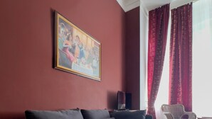 A modern living room with a dark sofa, a framed painting, a wicker chair, and a window with red curtains. - Hotel Maison Am Olivaer Platz (Berlin)