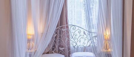 A four-poster canopy bed with white linens and towels, two bedside lamps, and a wicker chair.
