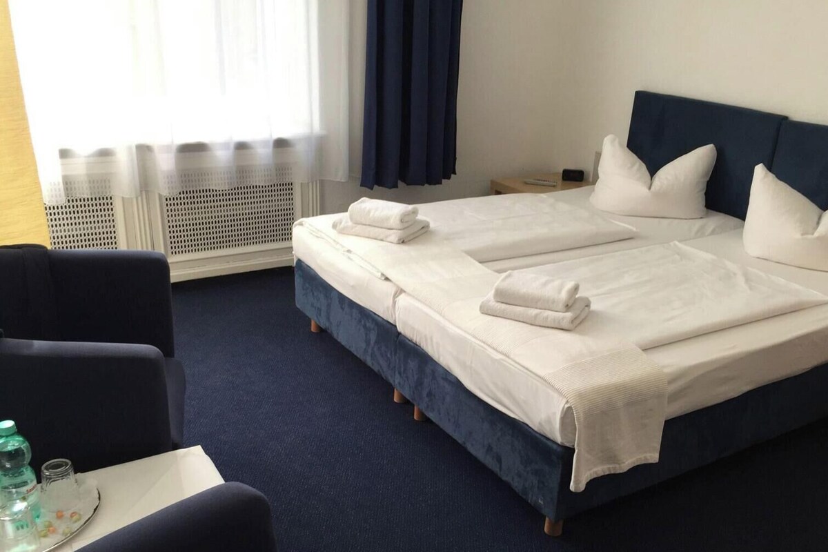 A hotel room with a double bed, two pillows, a bedside table, a chair, and a window with curtains.